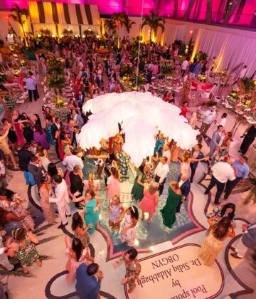 High-angle view of a crowded ballroom reception with round tables, a central floral display, and guests dancing on a circular floor inlay.