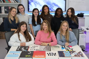 A group of nine young women and a teacher pose behind a table covered with various book covers and yearbooks in a classroom.