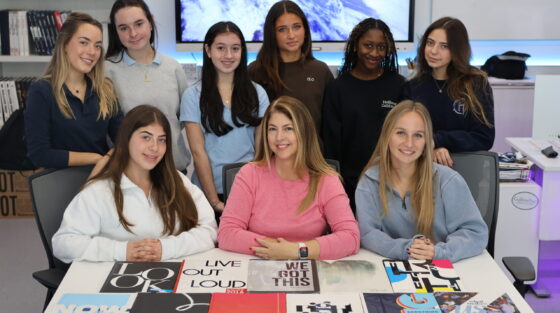 A group of nine young women and a teacher pose behind a table covered with various book covers and yearbooks in a classroom.