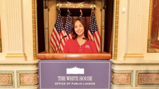A woman standing at a podium labeled “The White House Office of Public Liaison,” with two American flags behind her.