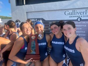Four members of a girls’ water polo team in navy blue uniforms hold a championship trophy in front of a Gulliver Prep swimming scoreboard.