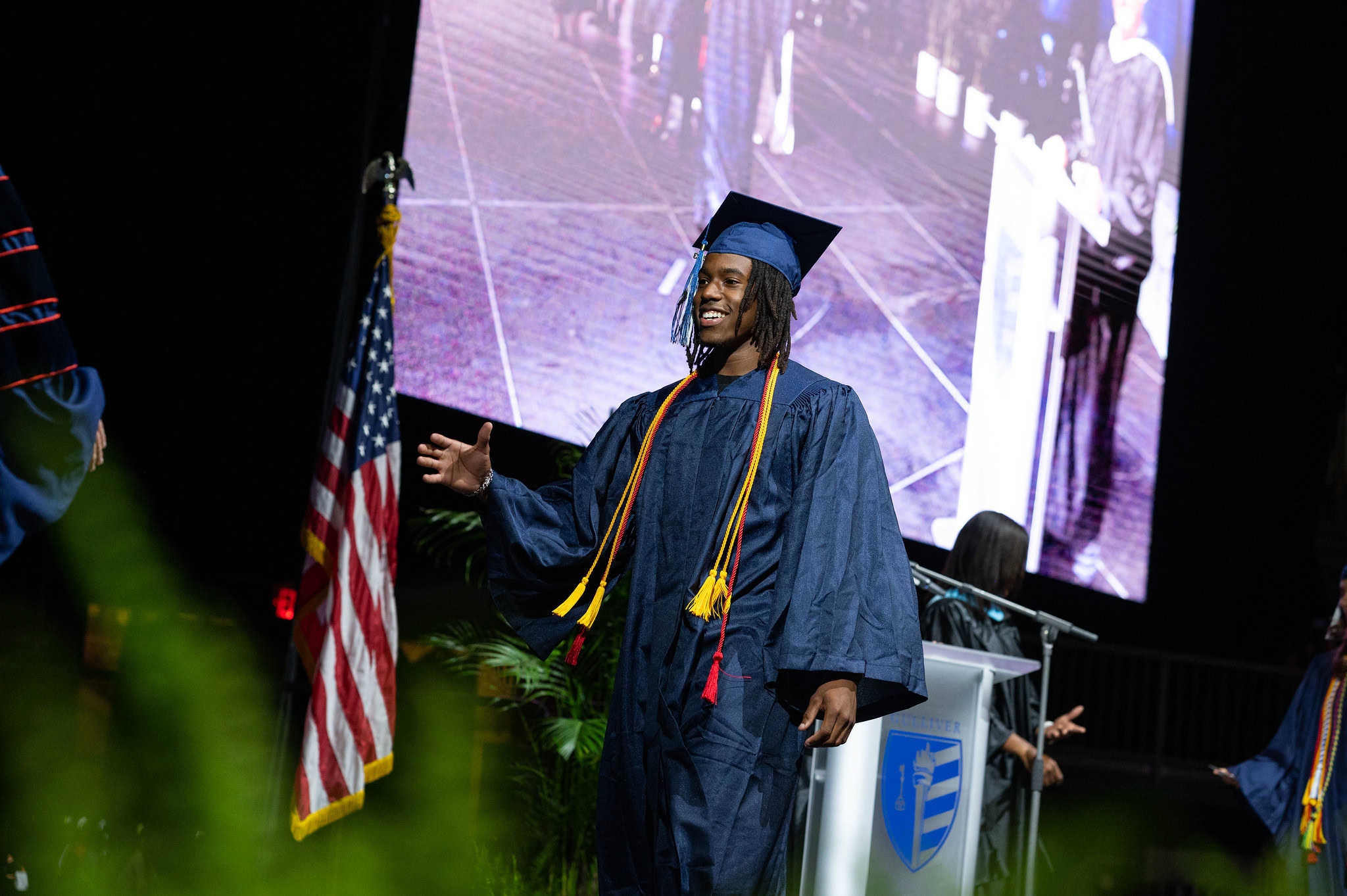 A graduate in a blue cap and gown walks across the stage with an outstretched arm at a graduation ceremony, with an American flag and a large screen in the background.