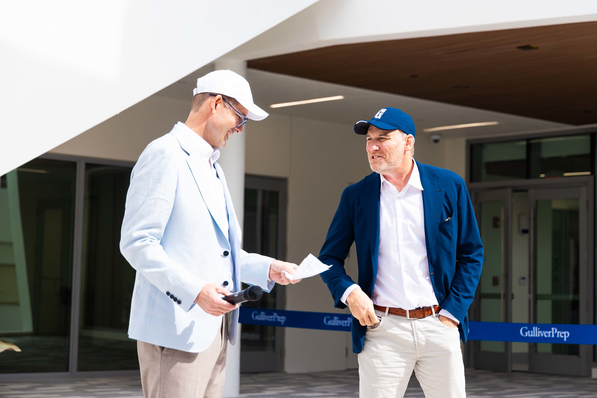 Two men in blazers, one holding papers, stand and talk outside a building with a “Gulliver Prep” sign visible in the background.