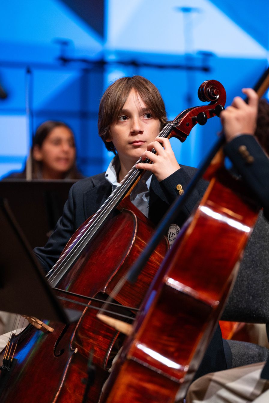 A young musician in formal attire plays the cello during a concert, with other performers and instruments visible in the background.