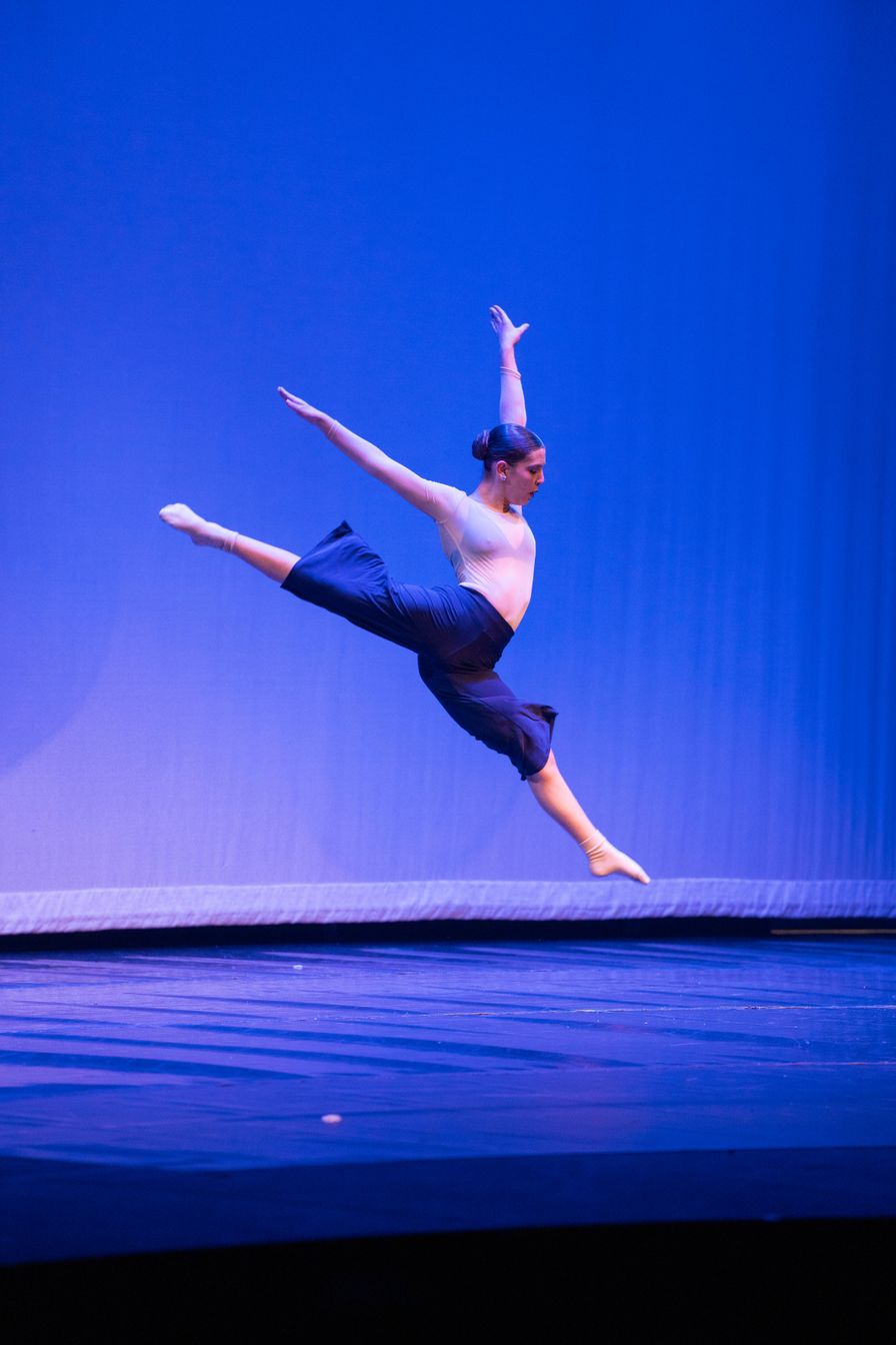 A dancer in a blue-lit theater leaps with arms and legs extended mid-air, wearing a light top and dark pants on a stage.