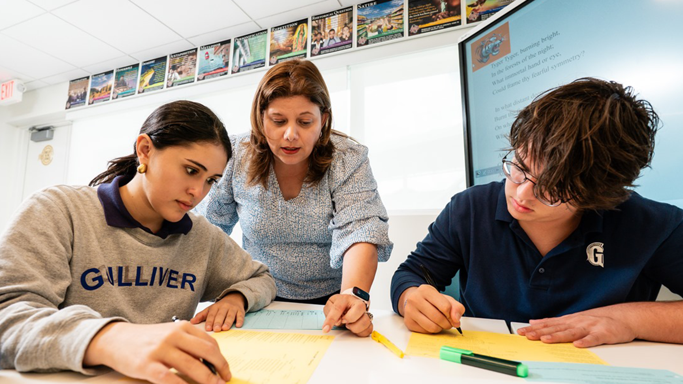A teacher assists two students working on assignments at a desk in a classroom. The students write on yellow sheets of paper. Educational posters are displayed on the walls.