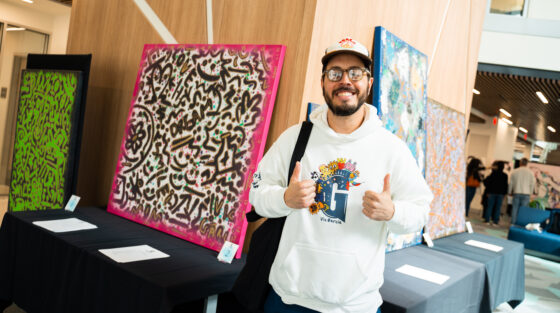 An artist wearing a white hoodie and baseball cap gives a thumbs-up in front of his abstract paintings for an alumni spotlight.