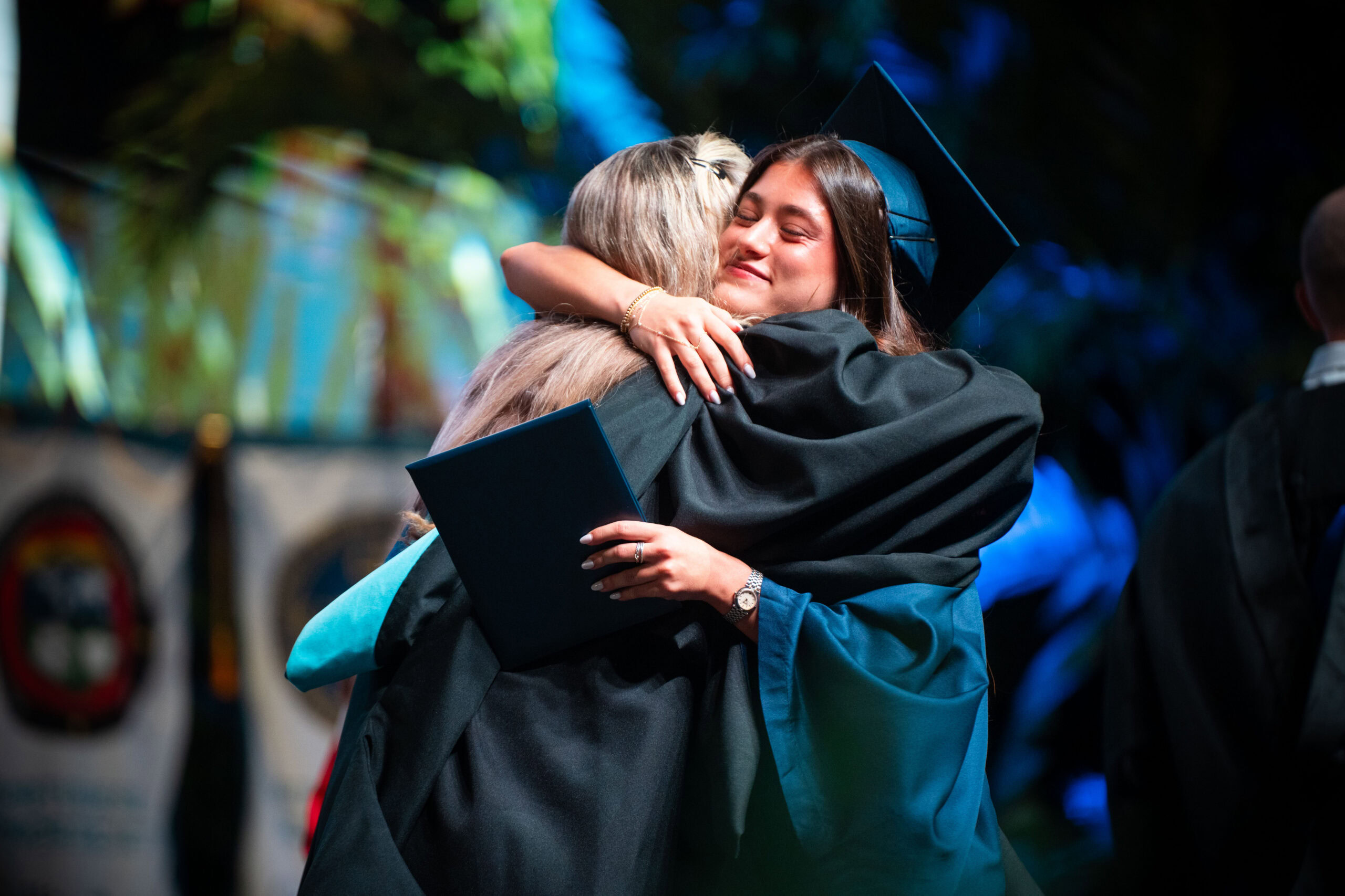 A graduate in a cap and gown embraces another person during a commencement ceremony.