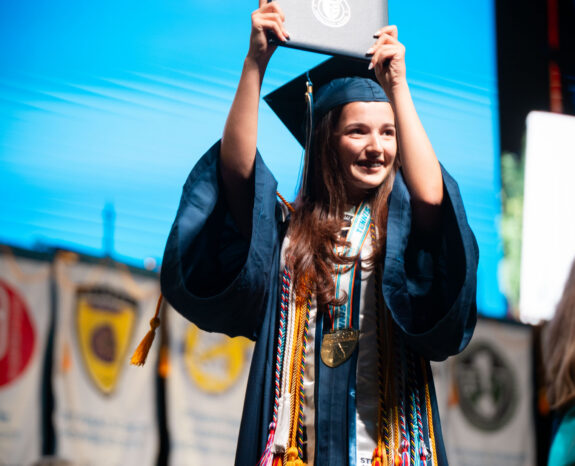 A graduate in cap and gown holds up a diploma on stage, wearing multiple honor cords and medals. Banners hang in the background.