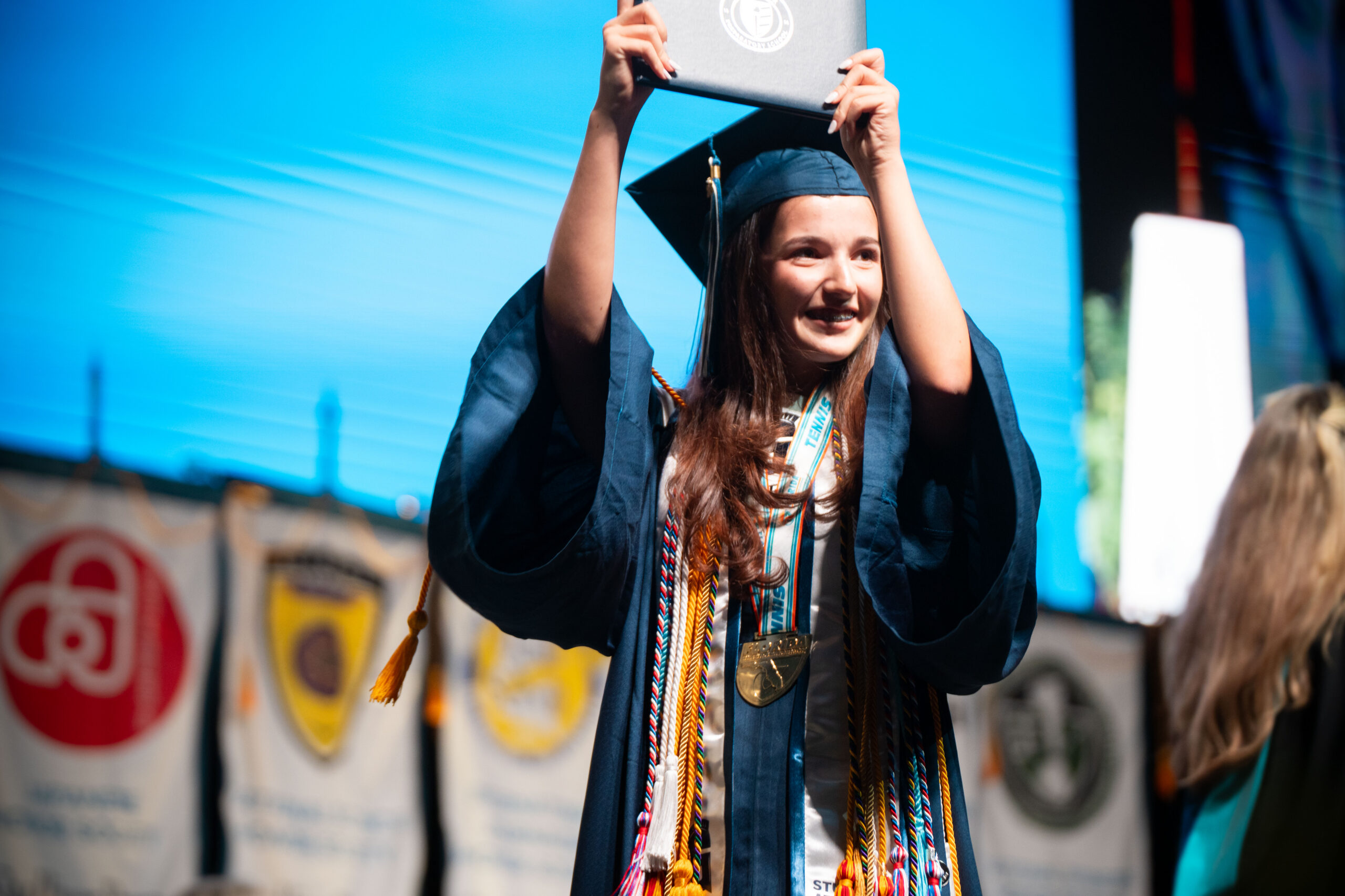 A graduate in cap and gown holds up a diploma on stage, wearing multiple honor cords and medals. Banners hang in the background.