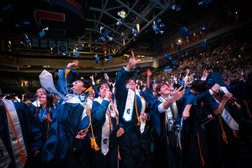 Graduates in caps and gowns cheer and throw caps into the air during a graduation ceremony in a crowded indoor arena.