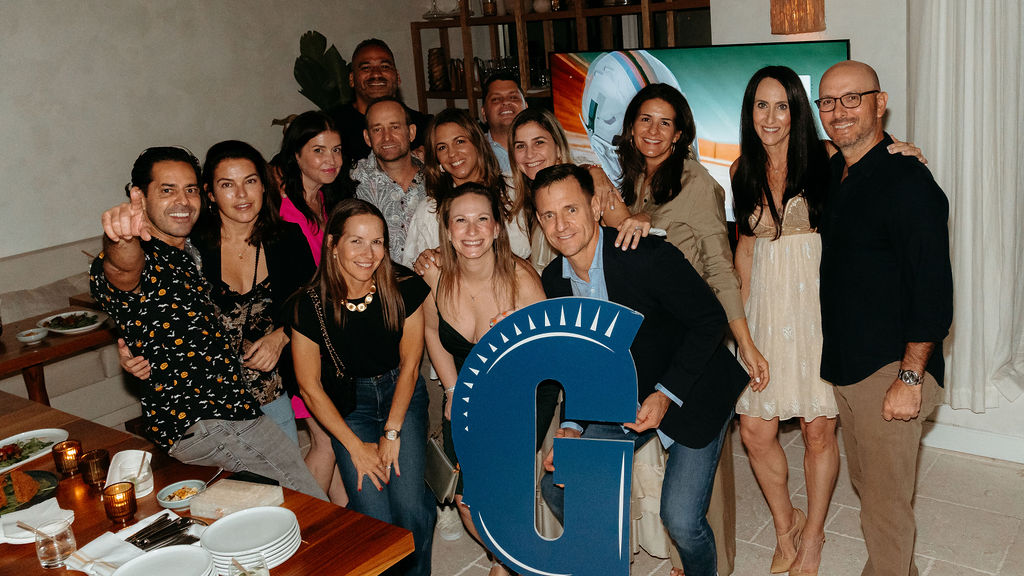 A group of people pose indoors at a gathering, some smiling at the camera. One person holds a large blue letter “G.” Food and drinks are visible on the table in the foreground.