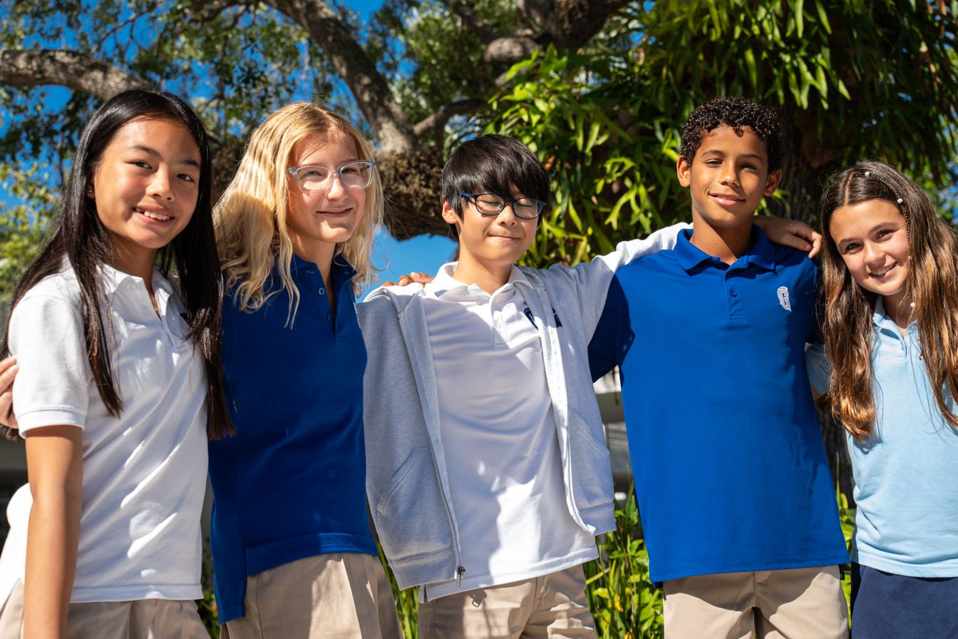 Five students wearing school uniforms stand outdoors with their arms around each other, smiling at the camera, with trees and greenery in the background.