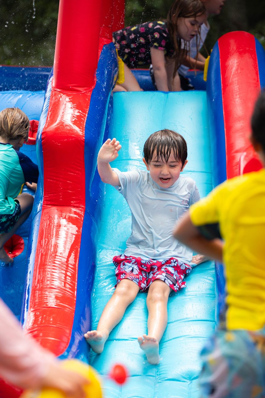 Young child slides down a wet, inflatable slide, raising one arm, while other children play nearby; everyone appears to be enjoying a water activity outdoors.