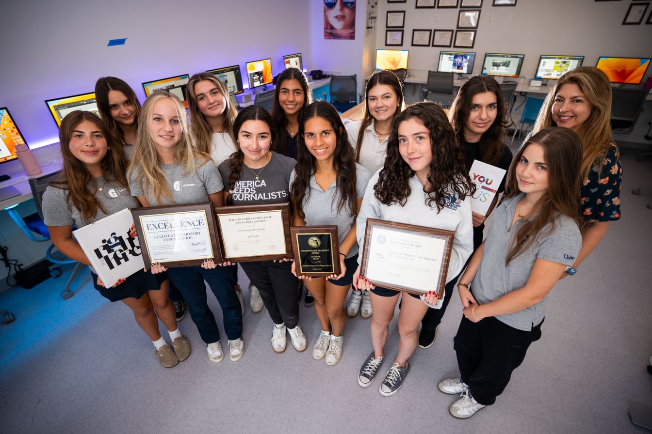 A group of students and a teacher pose in a computer lab, with some students holding journalism awards and certificates.