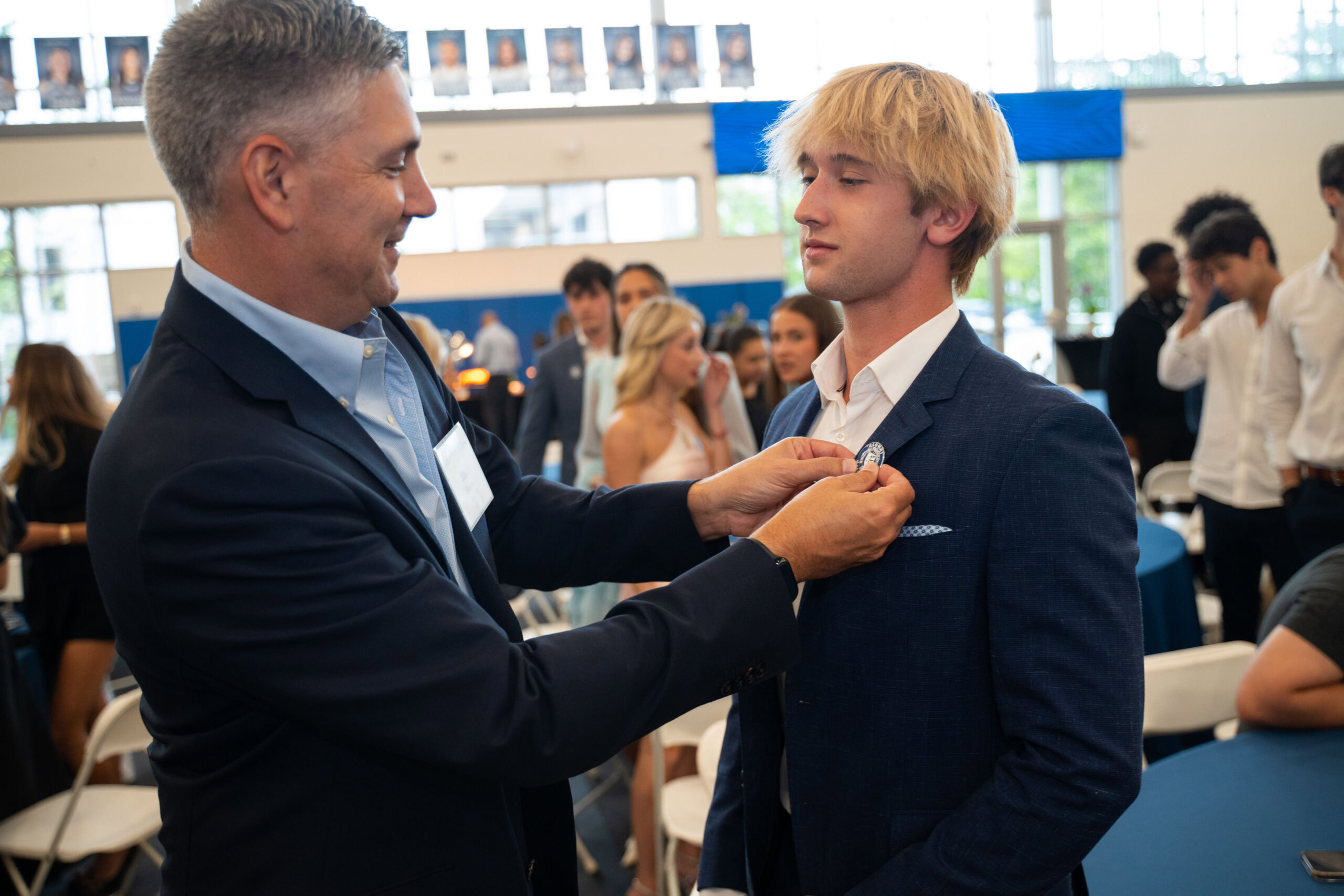 An older man pins a medal on a young man’s suit jacket in a crowded indoor event space with people in the background.