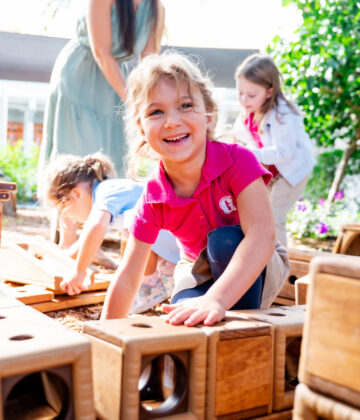 Four young children play outdoors, building with large wooden blocks. One girl in a pink shirt smiles at the camera while others focus on stacking and arranging the blocks.