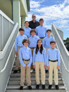 An adult man and eight children in light blue button-down shirts and khaki pants pose on an outdoor staircase.