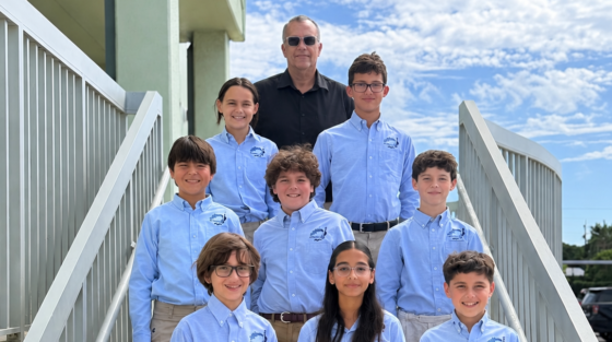 An adult man and eight children in light blue button-down shirts and khaki pants pose on an outdoor staircase.