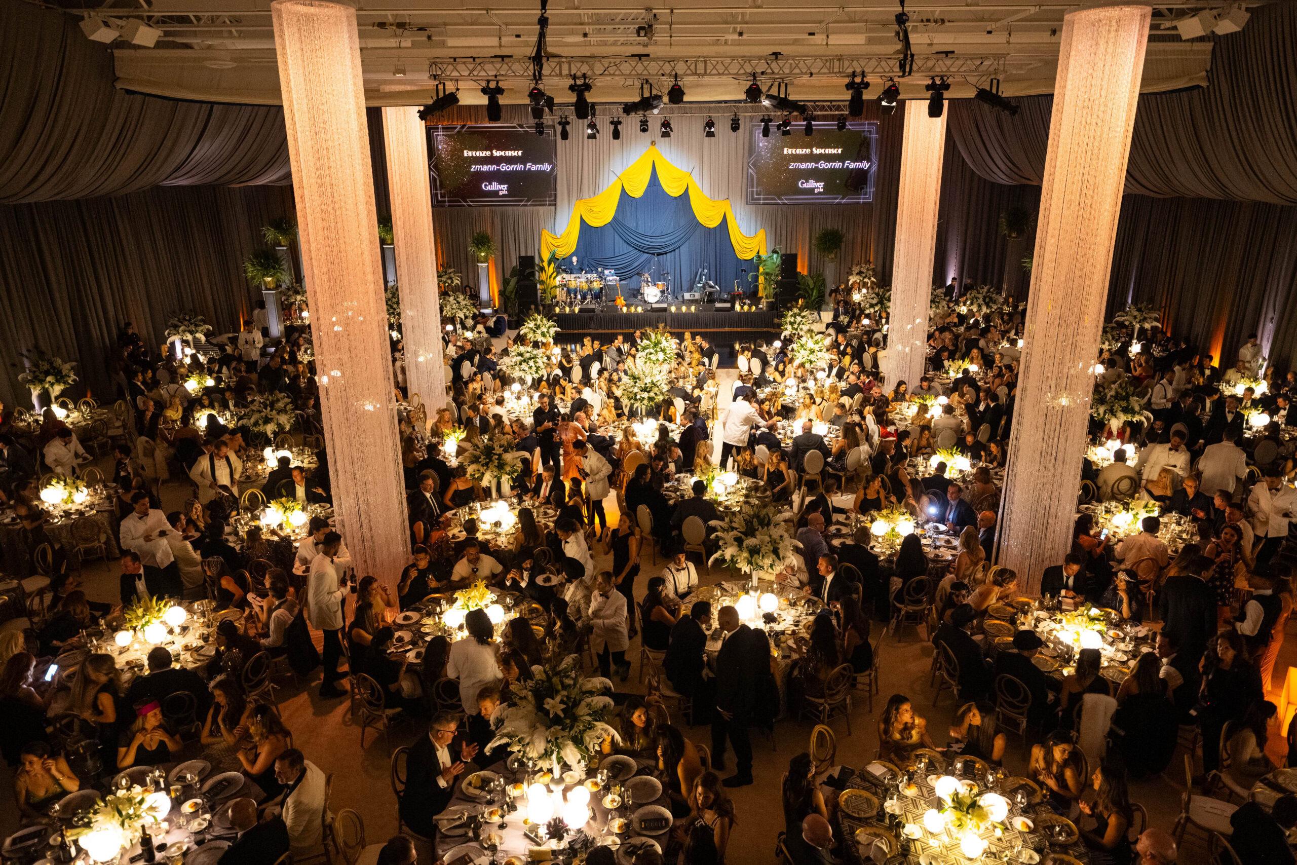 A large formal banquet with round tables, elegant lighting, and guests dressed in evening attire, centered around a stage with yellow and blue drapery and a live band setup.