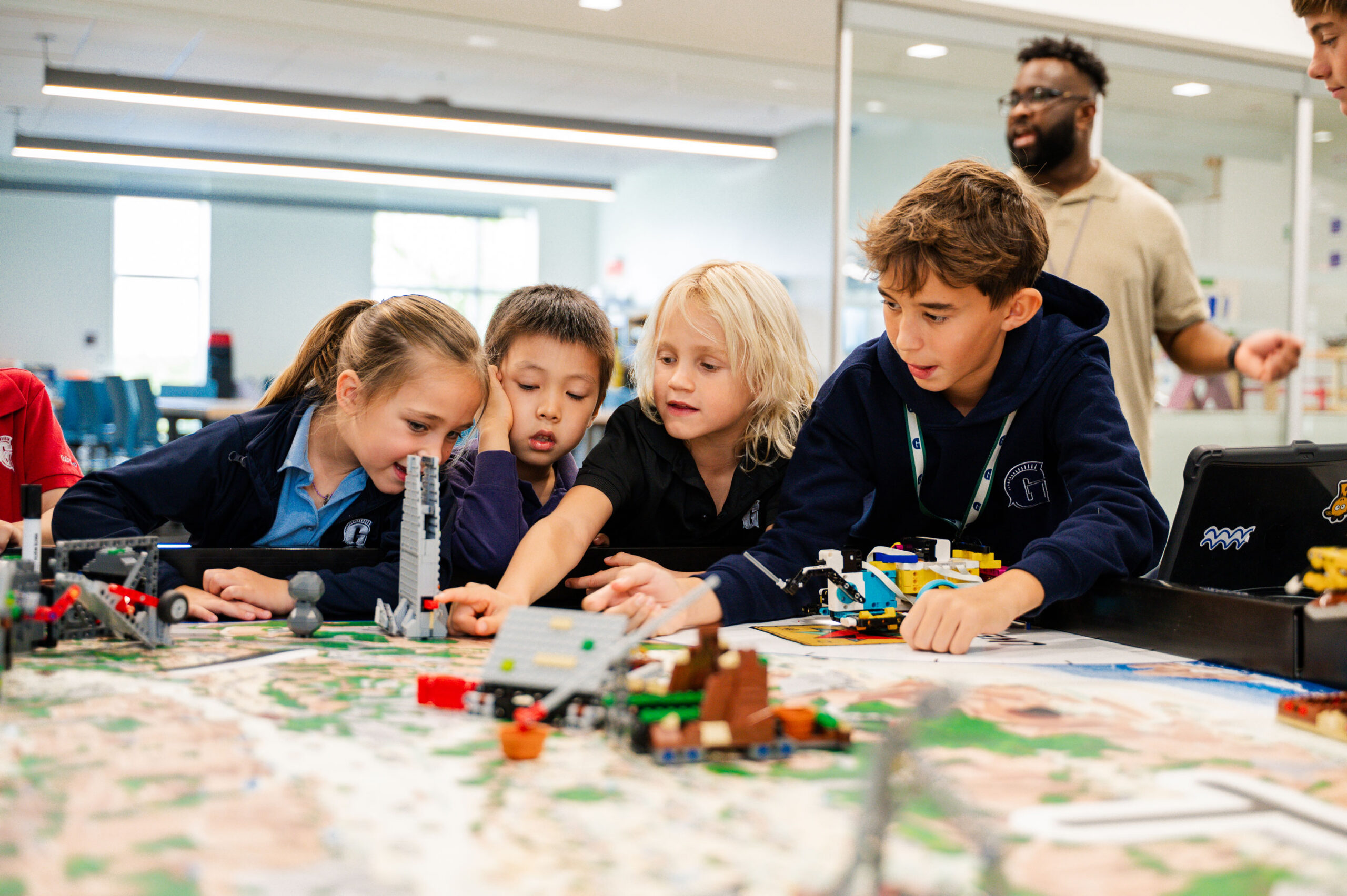 Four children work together on a robotics project with LEGO pieces at a table, while an adult stands in the background observing them.