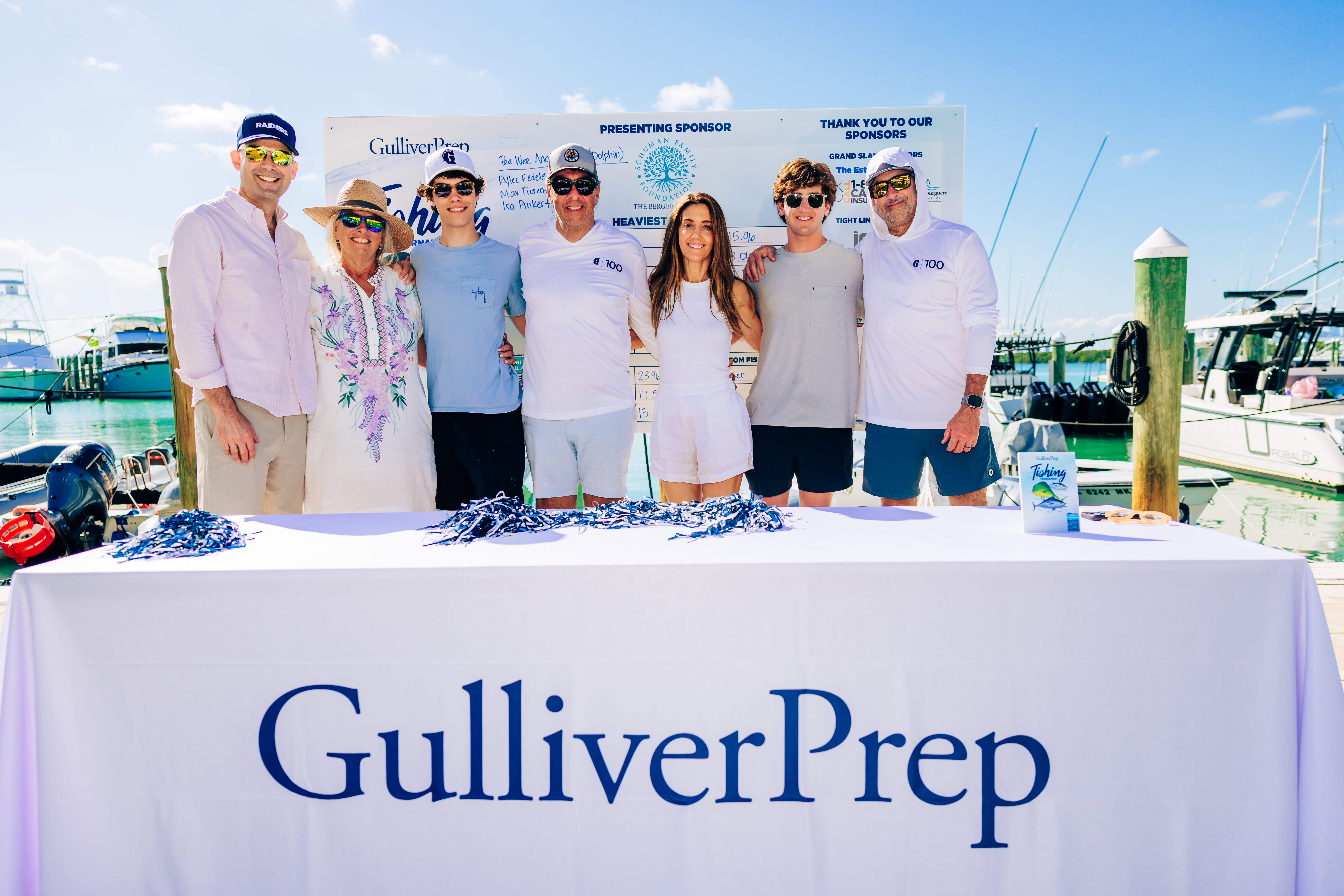 Seven people stand behind a table with a Gulliver Prep tablecloth at a marina, with boats and sponsor signs visible in the background.
