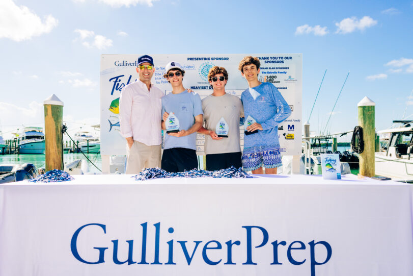 Four people stand behind a table with a “Gulliver Prep” sign, holding trophies, with boats and a sponsor board in the background under a clear sky.