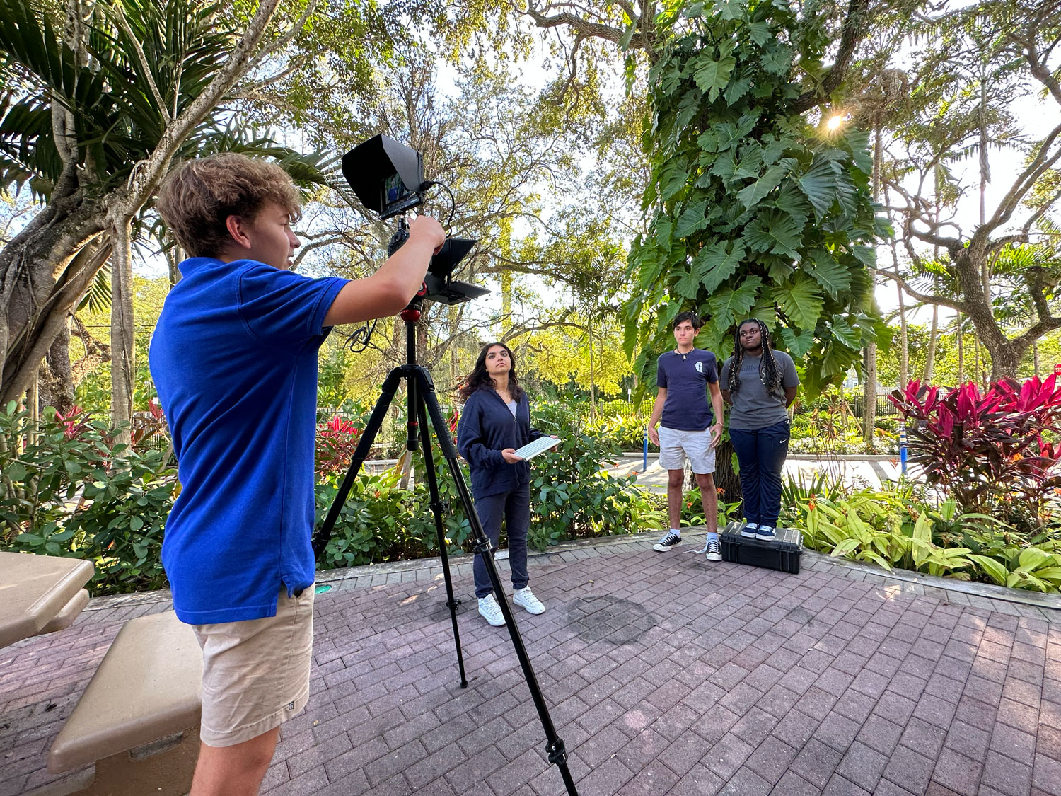 Four people are filming outdoors on a paved area. One person operates a camera on a tripod while the others stand in front of it, surrounded by trees and plants.