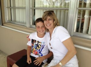 A woman and a young boy sit close together on a bench outside, smiling at the camera in this alumni spotlight. The boy wears a colorful shirt; the woman wears a white shirt and sunglasses on her head.