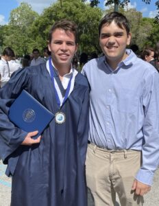 Two young men stand outdoors; one wears a graduation gown, medal, and holds a diploma folder in this alumni spotlight, while the other is in a light blue shirt.