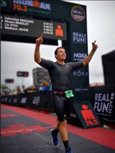 Alumni spotlight: Athlete crosses the finish line of an Ironman 70.3 Gulf Coast race, raising both hands in celebration, with race banners and timing display in the background.