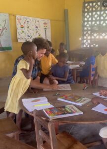 An alumni spotlight: A teacher helps two young students with their work at a round table, while other children are seated at desks and books and papers are spread out on the table.