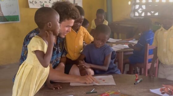 An alumni spotlight: A teacher helps two young students with their work at a round table, while other children are seated at desks and books and papers are spread out on the table.