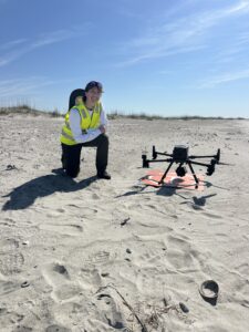 An alumni spotlight features a person in a high-visibility vest kneeling on a sandy beach next to a drone on a landing pad.