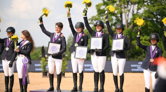 Five equestrians in riding gear stand on a podium holding sunflowers, medals, and award plaques.