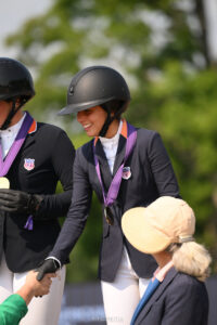 An equestrian athlete in a helmet and competition jacket wearing a medal shakes hands with a person in a sun hat.