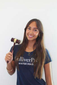 A smiling young woman in a Gulliver Prep Water Polo t-shirt holds up a wooden gavel.