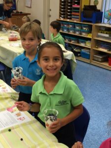 Two elementary students in school uniforms hold cups of yogurt and blueberries in a classroom.