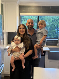 An alumni spotlight showing a man and woman standing in a kitchen, holding a baby and a toddler.