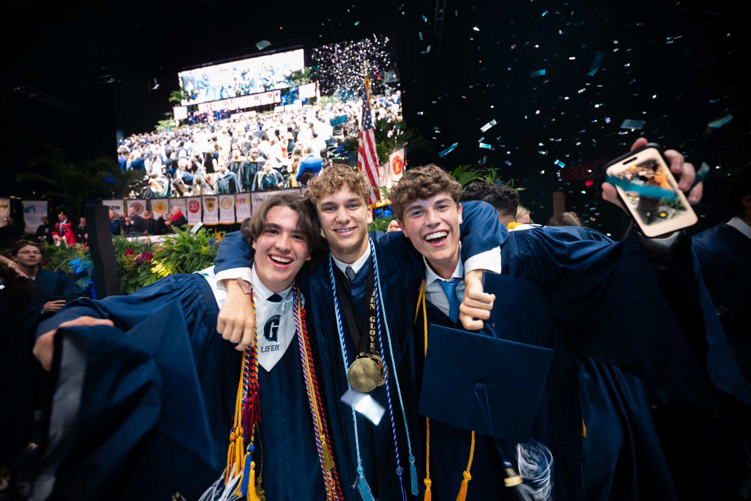 Three graduates in blue gowns and honor cords smile and pose together, holding diplomas, with confetti falling and a crowd celebrating in the background.