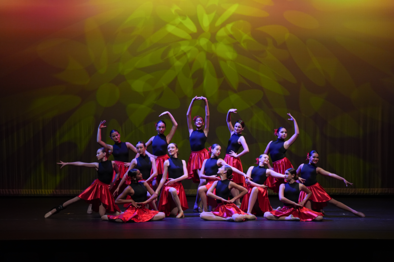 A group of dancers in black tops and red skirts pose on stage, some standing and some seated, under yellow stage lighting with a patterned backdrop.