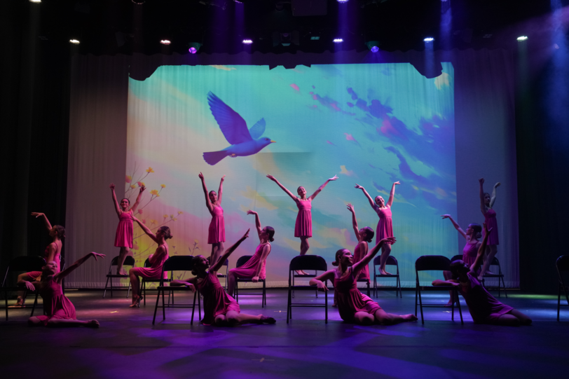 A group of dancers in pink costumes perform on stage with chairs, arms raised, against a backdrop of a flying bird and sky.