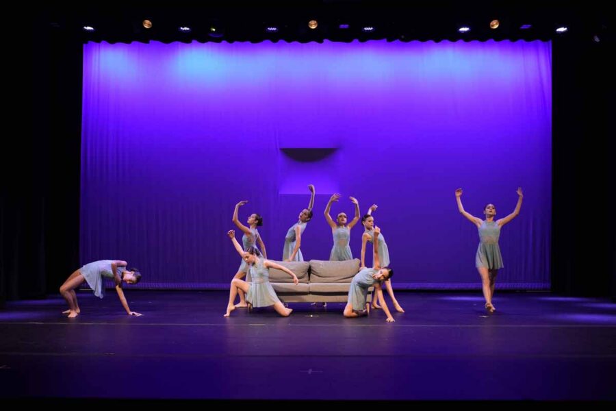 Eight dancers in light blue costumes perform on stage around a beige sofa, striking expressive poses against a purple-lit backdrop.