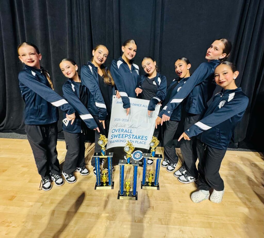 Eight girls in matching athletic jackets pose together onstage, smiling and gesturing toward several trophies and a large sign that reads “Overall Sweepstakes, Ranking 1st Place.”.