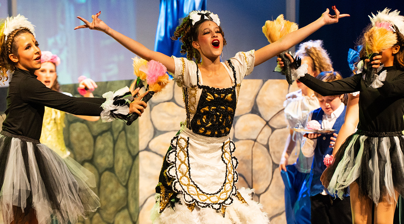A group of children in colorful costumes perform on stage; one girl in a white dress stands center with arms outstretched, while others hold feather dusters around her.