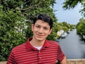A young man in a red striped polo shirt smiling on a bridge overlooking a canal with boats.