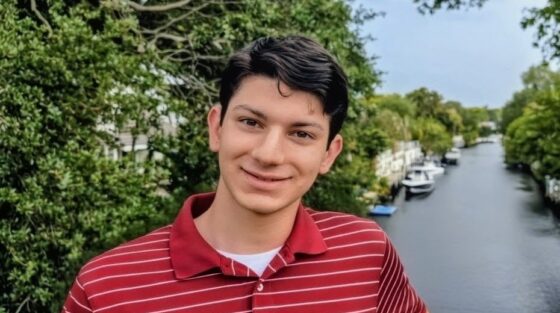 A young man in a red striped polo shirt smiling on a bridge overlooking a canal with boats.