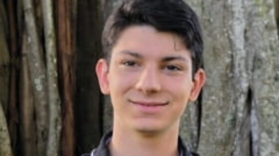 A young person with short dark hair wearing a black collared shirt stands in front of a large tree trunk, smiling at the camera.