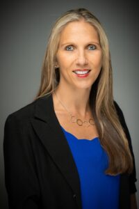 Professional headshot of a woman with long blonde hair, wearing a black blazer over a blue top and a geometric necklace.