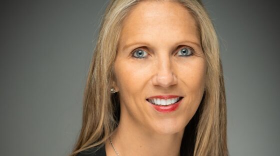 Professional headshot of a woman with long blonde hair, wearing a black blazer over a blue top and a geometric necklace.